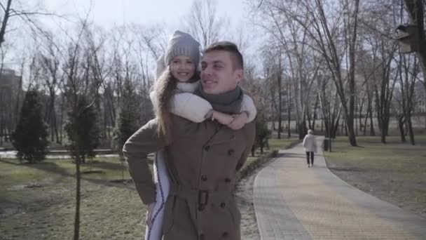 Портрет щасливого кавказького молодого батька, який тримає маленьку гарну дочку на спині. Весела дівчина і чоловік стоять у сонячному осінньому парку і посміхаються. Життя, сім'я, щастя, єдність. — стокове відео