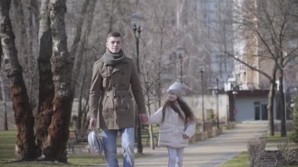 Retrato de una niña caucásica positiva caminando desde la escuela con su padre. Hombre joven sosteniendo niños mochila y paseando con su hija en el parque de otoño. Familia, responsabilidad, estilo de vida . — Vídeos de Stock