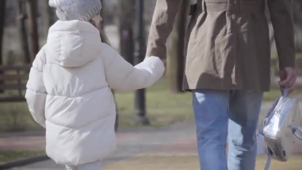 Achteraanzicht van klein schoolmeisje wandelend met jonge vader langs steegje in de herfst of lente park. Een meisje in een witte jas die loopt na een studie met haar ouders. Lifestyle, familie, ondersteuning. — Stockvideo