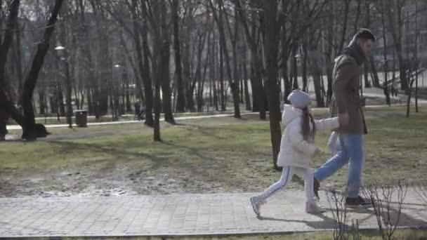 A fiatal kaukázusi apa oldalnézete arra kényszeríti a kislányát, hogy iskolába járjon. Vonakodó aranyos iskoláslány sétál az iskolába apával elhaladó őszi vagy tavaszi parkban. Életmód, tanulmányok, család. — Stock videók