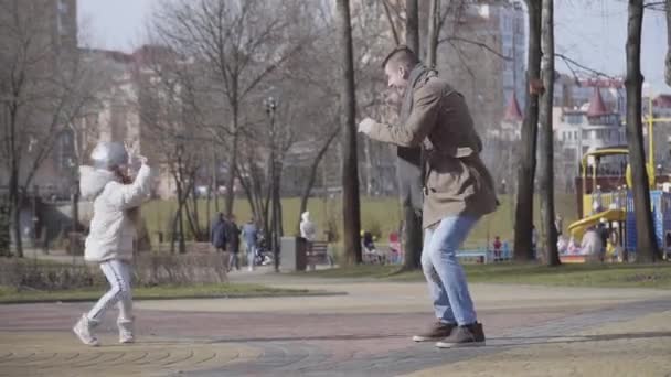 Alegre padre joven caucásico e hija pequeña bailando en el soleado parque de otoño. Vista lateral de hombre y chica sonrientes divirtiéndose al aire libre. Estilo de vida, alegría, felicidad, ocio . — Vídeo de stock