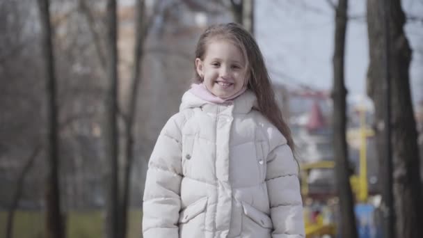 Портрет радостной маленькой девочки, сидящей перед камерой осенью или в весеннем парке. Счастливый улыбающийся кавказский ребенок веселится на улице по выходным. Радость, счастье, досуг, образ жизни . — стоковое видео