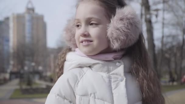 Gelukkig meisje in oorkappen genietend van de zonnige dag in het park. Portret van een schattig blank kind dat in de buitenlucht rondkijkt. Lifestyle, vrije tijd, geluk, jeugd. — Stockvideo