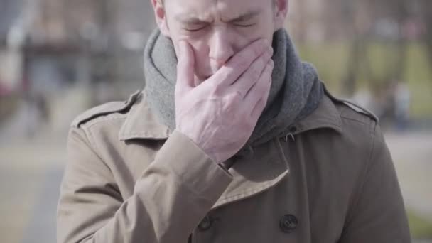 Close-up van de jonge zieke man die op de bank zit en de keel wrijft. Portret van een zieke blanke man buiten op zonnige herfst- of lentedag. Gezondheid zijn, ziekte, verkoudheid en griep, ademhalingsziekte. — Stockvideo