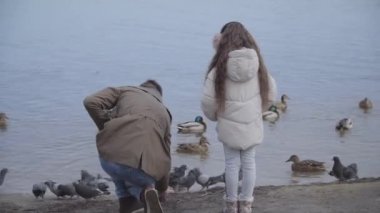 Uzun kıvırcık saçlı esmer kızın nehir kıyısında dikilip babalarının ördek yemlerine bakışı. Çocuk ve ebeveyn bahar ya da sonbahar günü dışarıda dinleniyorlar. Birlik, aile, eğlence, yaşam tarzı.