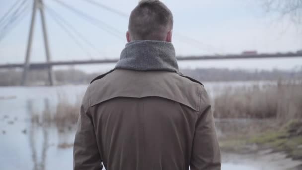 Baksidan av ung brunett vit man promenader längs floden. Kamera följer omtänksam stilig kille promenader ensam utomhus. Ensamhet, livsstil, natur, fritid. — Stockvideo