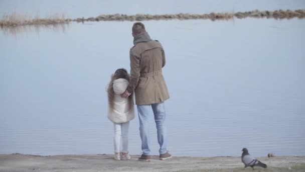 Achteraanzicht van de jonge blanke vader die met zijn dochter op de oever van de rivier staat te praten. Breed beeld van een man en een klein meisje die buiten kletsen op de achtergrond van rustig water. Landschap, natuur, vrije tijd. — Stockvideo