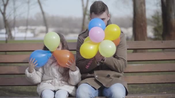 Позитивний молодий чоловік і маленька дівчинка рухаються по кольорових повітряних кулях і посміхаються перед камерою. Весела кавказька дочка і батько, що полювали на лавку в осінньому парку. дозвілля, радість, спосіб життя, щастя. — стокове відео