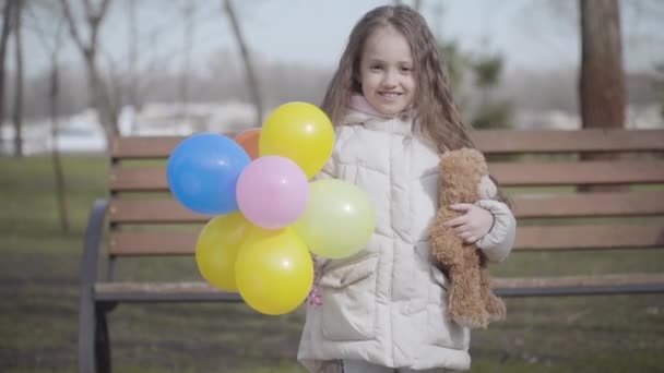 Happy brunette Caucasian girl posing outdoors with balloons and teddy bear. Portrait of smiling kid with brown eyes enjoying sunny day in park. Joy, leisure, happiness, childhood, lifestyle. — Stock Video