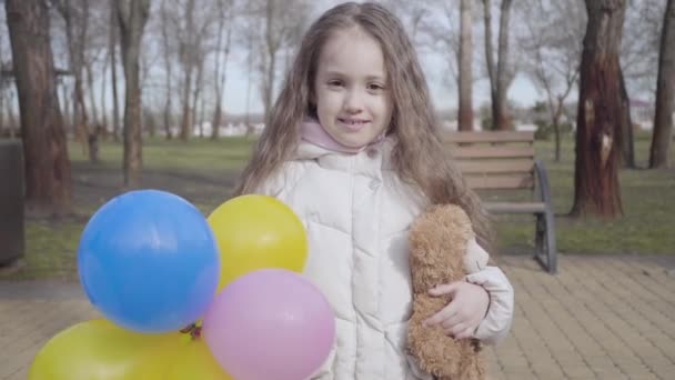 Porträt eines kleinen fröhlichen Kindes, das im sonnigen Frühling- oder Herbstpark posiert. Brünettes kaukasisches Mädchen mit lockigem Haar, Teddybär und Luftballons in der Hand und lächelnd in die Kamera. Freude, Muße, Glück, Genuss. — Stockvideo