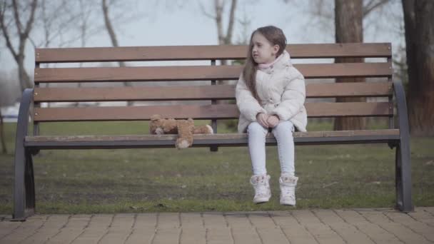 Crying lost little girl rubbing eyes as sitting on bench in spring or autumn park. Portrait of pretty lonely Caucasian kid with teddy bear. Childhood, sadness, social problem, anxiety. — Stock Video