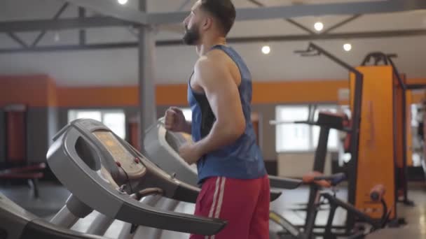 Spor kulübünde koşu bandında koşan atletik Orta Doğulu bir adamın yan görüntüsü. İçeride antrenman yapan güçlü bir sporcu. Sağlıklı yaşam tarzı, spor, atletizm, koşu, spor. — Stok video