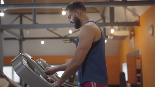 スポーツ中東の男は、トレッドミルやジョギングに速度を追加します。ジムでの健康的なスポーツマンのトレーニングの笑顔の側面図。健康、運動、持久力. — ストック動画