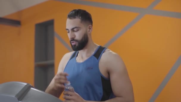 Retrato del deportista corriendo limpiando la frente con la mano y el agua potable. Joven hombre confiado de Oriente Medio terminando el entrenamiento en la cinta de correr en el gimnasio. Estilo de vida, atletismo, entrenamiento . — Vídeo de stock