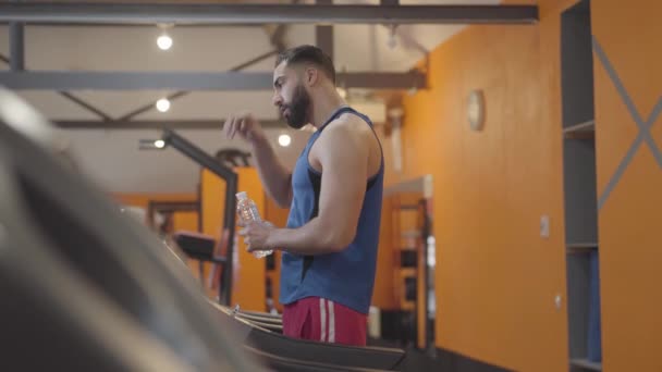 Sidovy av utmattad Mellanöstern idrottsman torka pannan och dricksvatten efter träning. stilig man med svart hår som tränar på löpband i gymmet. Sport, träning, hälsosam livsstil. — Stockvideo