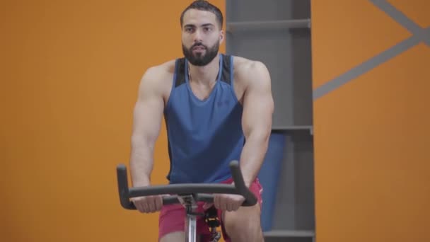 Retrato de un hombre concentrado de Oriente Medio montando bicicleta estática en el gimnasio. Deportista fuerte y seguro que ejerce en el club deportivo. Fitness, deporte, masculinidad, estilo de vida saludable . — Vídeos de Stock