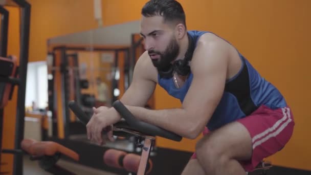 Primer plano del deportista exhausto pedaleando bicicleta estática. Vista lateral del cansado joven Oriente Medio equipo de equitación en el gimnasio. Salud, estilo de vida, deporte, resistencia, entrenamiento . — Vídeo de stock