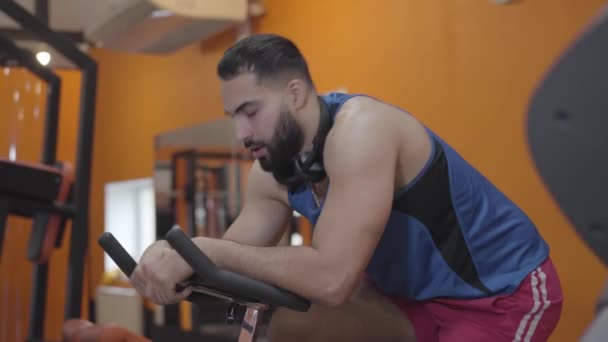 Vista lateral del deportista cansado limpiando la frente y suspirando. Joven hombre de Oriente Medio exhausto montar bicicleta estática en el club deportivo. Estilo de vida saludable, deporte, resistencia, entrenamiento . — Vídeo de stock