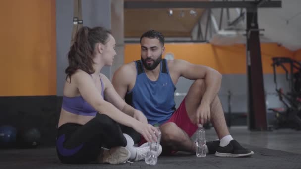 Joven hermosa mujer caucásica y guapo hombre de Oriente Medio charlando en el gimnasio y el agua potable. Deportista delgado fuerte y deportista descansando después del entrenamiento en el club deportivo. Estilo de vida saludable . — Vídeos de Stock