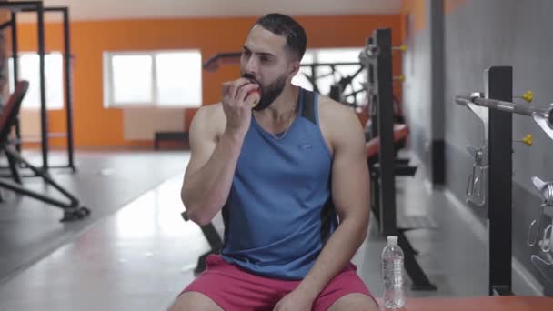 Um tiro no meio de um homem musculoso do Médio Oriente a comer maçã no ginásio. Retrato de um belo jovem desportista a fazer uma pausa no treino. Estilo de vida, confiança, alimentação saudável, esporte . — Vídeo de Stock