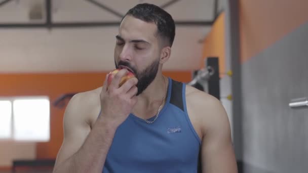 Primer plano del apuesto deportista de Oriente Medio con ojos marrones mordiendo manzana roja y masticando. Retrato de hombre feliz confiado comiendo fruta después del entrenamiento. Deporte, estilo de vida, salud . — Vídeo de stock