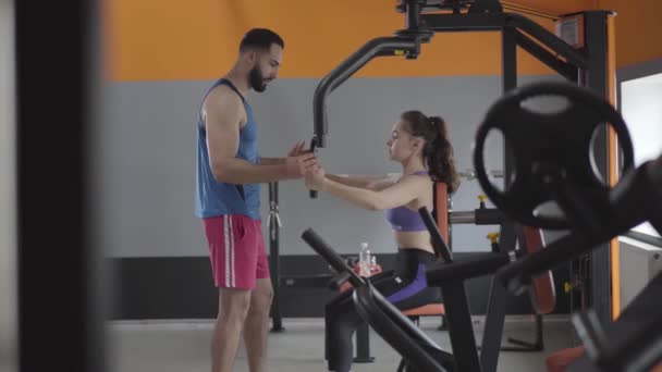 Amplio tiro de mujer caucásica joven delgada entrenar los músculos de la mano y escuchar los consejos de entrenador personal. Vista lateral de hermosa chica morena y guapo hombre de Oriente Medio en el gimnasio . — Vídeos de Stock