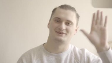 Genç esmer adam kameraya selam veriyor ve gülümsüyor. Video sohbetinde birini selamlayan neşeli beyaz adamın portresi. Uzaktan çalışma, konferans, Covid-19 yaşam tarzı. S-seyir defteri 2, QuickTime ProRes 422 HQ