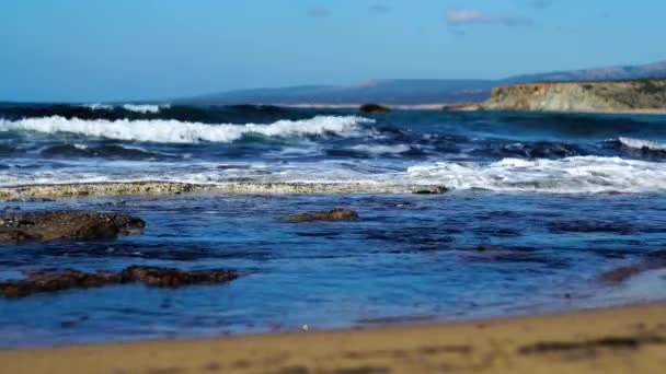 Ciemnoniebieskie fale morskie pluskające się na piaszczystej plaży w słoneczny letni dzień. Piankowe wody Morza Śródziemnego rozbijają się o skały i toczą się po piasku. Tropikalny raj, piękno natury, krajobraz morski. — Wideo stockowe