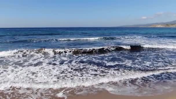 Kamera se pohybuje podél písečné letní pláže s pěnivými tmavomodrými vlnami. Slunečný den na majestátní kyperské turistické letovisko. Krásné vody Středozemního moře šplouchající na slunci. — Stock video