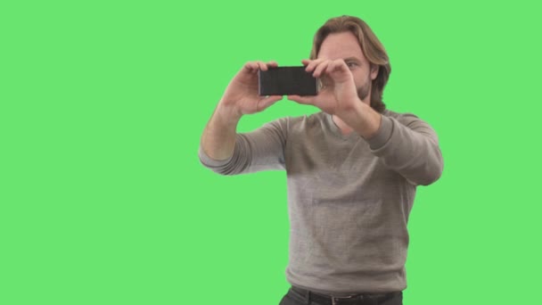 Μελαχρινή νεαρός άνδρας λήψη φωτογραφιών στο smartphone. Πορτρέτο του Καυκάσου φωτογράφου χρησιμοποιώντας συσκευή σε πράσινο φόντο. Τρόπος ζωής, χόμπι, δημιουργικότητα. Χρώμιο, πράσινη οθόνη. — Αρχείο Βίντεο