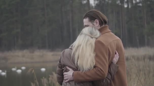 Vista trasera de la feliz pareja adulta abrazándose frente al lago con cisnes y bosque al fondo. Amoroso hombre y mujer caucásicos pasar nublado día de primavera u otoño al aire libre. Amor, ocio, citas. — Vídeo de stock