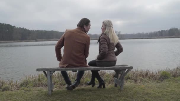 Visão traseira de homem e mulher positivos sentados no banco em frente ao lago da floresta e conversando. Amplo tiro de casal feliz amoroso caucasiano descansando ao ar livre no dia nublado. Lazer, amor, casamento, estilo de vida . — Vídeo de Stock
