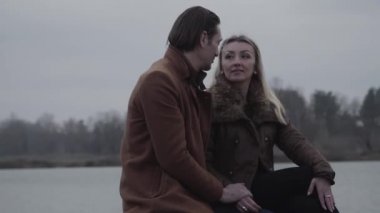 Bulutlu bir günde göl kenarında oturup konuşan mutlu çiftin orta pozu. Yakışıklı, beyaz tenli esmer bir adam ve güzel sarışın bir kadın açık havada çıkıyorlar. Boş zaman, yaşam tarzı, aşk, birliktelik..
