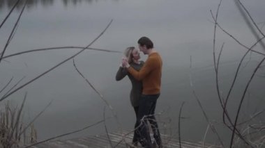 Ahşap iskelede aşk dolu beyaz bir çiftin romantik dansı. Bulutlu bir günde gölün kenarında dans eden ve gülümseyen mutlu erkek ve kadının geniş açılı görüntüsü. Aşk, romantizm, eğlence, bağ..