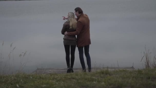 Hombre caucásico confiado señalando y hablando con la mujer rubia feliz como de pie en el muelle en la orilla del lago. Vista posterior amplia toma de pareja positiva para adultos citas al aire libre. Ocio, relajación, amor . — Vídeos de Stock