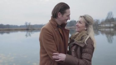 Gülümseyen yetişkin beyaz bir çiftin göl kıyısında kucaklaşması ve yürümesi. Dışarıda gezen mutlu karı kocanın portresi. Evlilik, aşk, flört, yaşam tarzı..