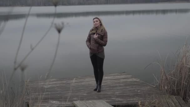 Mujer caucásica rubia pensativa de pie en el muelle de madera junto al lago. Amplio retrato de la hermosa dama encantadora pasar el día nublado solo al aire libre. Confianza, estilo de vida, soledad, belleza . — Vídeo de stock