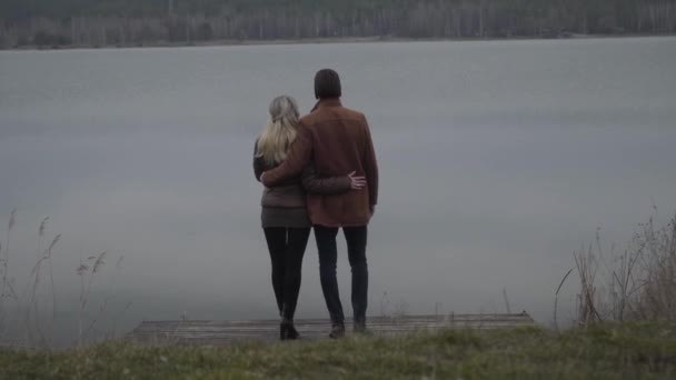 Wide shot of adult Caucasian man and woman standing on pier at lake shore and looking away. Back view of slim blond lady and brunette guy hugging outdoors. Love, dating, leisure, lifestyle. — Stock Video