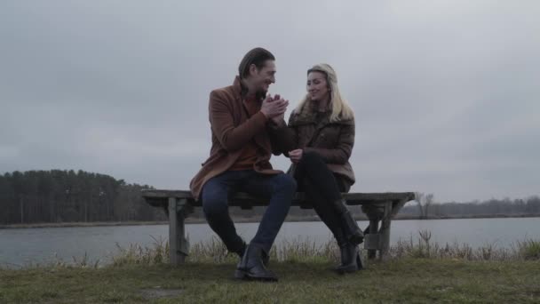 Šťastný milující muž hřející přítelkyně ruku s dechem. Široký portrét okouzlujícího bělošského páru sedícího na lavičce na břehu jezera a mluvícího. Láska, randění, volný čas, životní styl. — Stock video