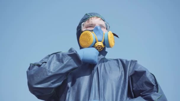Portrait de virologiste en costume de biodanger et respirateur posant à l'arrière-plan du ciel bleu clair d'été ou de printemps. Homme caucasien sérieux zippant costume chimique et regardant la caméra. Covid-19 . — Video