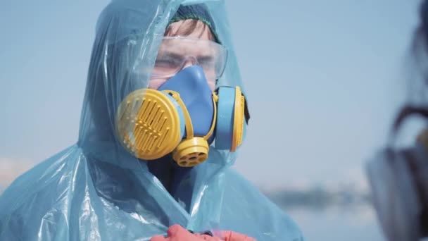 Люди одягаються один в одного в костюми біологічної небезпеки на відкритому повітрі. Портрет серйозного молодого кавказького чоловіка в респіраторі з допомогою колеги перед дезінфекцією. Коїд-19 пандемія, хімічне забруднення. — стокове відео