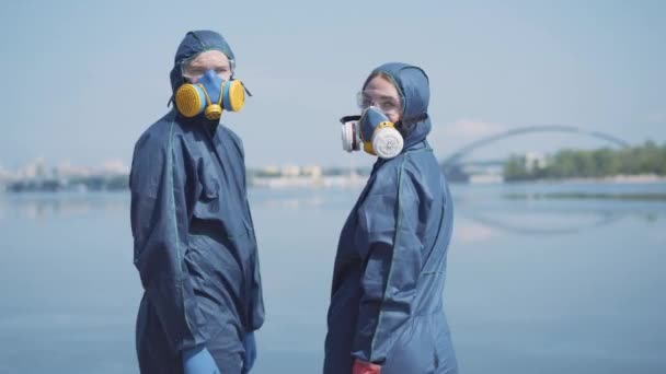 Mann und Frau in Atemschutzmasken und Chemikalienanzügen wenden sich der Kamera zu. In der Mitte ein Porträt junger kaukasischer Arbeiter, die nach der Desinfektion am Flussufer posieren. Covid-19-Pandemie, Lebensstil, Umweltverschmutzung. — Stockvideo