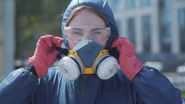 Gros plan portrait de jeune belle femme caucasienne en costume chimique et lunettes de protection portant un respirateur et regardant la caméra. Spécialiste féminine confiante posant à l'extérieur le jour de l'été . — Video