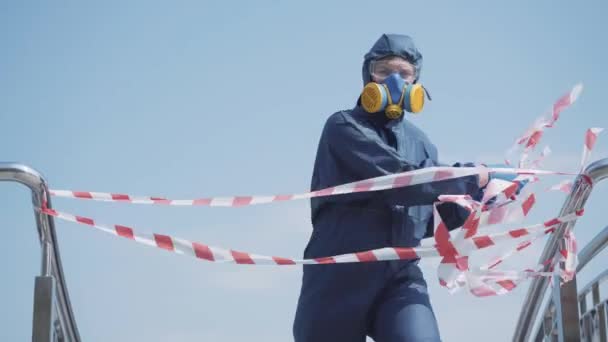 L'homme en costume chimique arrachant la bande de prudence et s'en va. Portrait de jeune homme caucasien en respirateur enlever la barrière d'avertissement à l'extérieur. Covid-19, risque biologique radioactif pollution atmosphérique . — Video