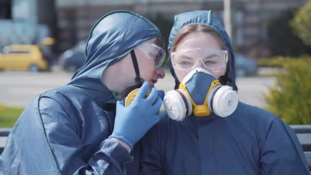 Detailní záběr mladého radostného muže šeptajícího na ucho krásné ženy v chemickém obleku a respirátoru. Legrační, usmívající se bělošský pár drbající venku. Covid-19, humor, zábava, životní styl. — Stock video