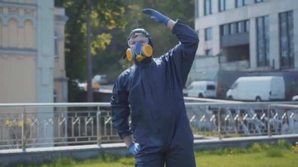 Zábavné cvičení muže v respirátoru a chemickém obleku venku. Portrét mladého kavkazského sportovce, který se protahuje za slunečného dne během pandemie Covid-19 nebo znečištění ovzduší. Sranda, humor, životní styl. — Stock video