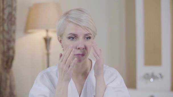 Belle femme aux yeux gris blonds touchant le visage et souriant. Portrait de dame blanche heureuse confiante en peignoir blanc appliquant crème visage anti-âge. Rajeunissement, hydratation, beauté, style de vie . — Video