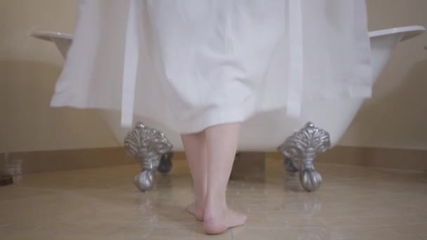 Peignoir blanc tombant, jambes féminines caucasiennes debout devant la baignoire. Vue arrière de la jeune femme méconnaissable prenant un bain à la maison. Détente, beauté, soins du corps, hygiène, style de vie . — Video