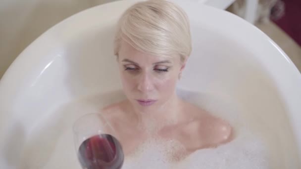 Vue de dessus gros plan de charmante femme blonde avec verre à vin regardant la caméra et souriant. Portrait de belle dame caucasienne relaxant dans la baignoire avec du vin rouge. Mode de vie, bonheur, détente, joie . — Video