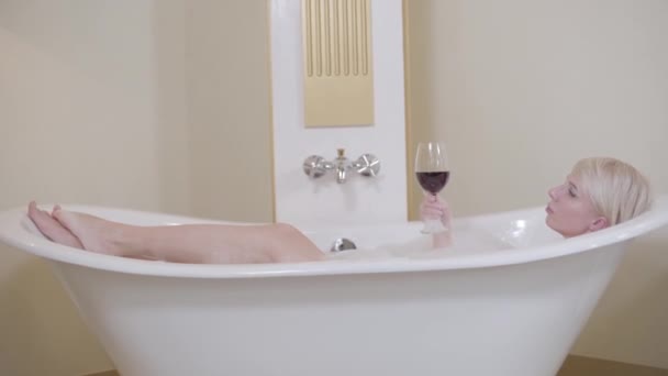 Mulher caucasiana loira confiante bebendo vinho tinto no banheiro. Vista lateral tiro largo de bela senhora adulta deitada no banho com espuma. Lazer da noite, estilo de vida, mimos, relaxamento, alegria . — Vídeo de Stock